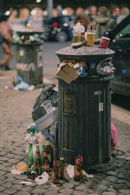 A black, cylindrical public rubbish bin made of plastic or metal with a domed top, situated on a cobblestone pavement in an urban area. The bin’s surface appears weathered, with visible scratches and marks. It is overflowing with various types of waste, including crumpled paper and plastic bags protruding from the openings. Surrounding the bin on the ground are numerous empty glass bottles, mainly green and brown, some broken or partially crushed, along with crumpled paper and plastic wrappers. On top of the bin, there are disposable cups with lids and straws, a paper food container, and a red and white disposable cup, indicating recent use. In the background, blurred figures of people are visible, suggesting a busy street scene, with additional parked cars and another rubbish bin further away. The overall scene depicts urban littering and collection issues, underscoring the importance of effective rubbish clearance and private waste management services, such as those offered by Rubbish Clearance Bayswater, particularly in busy public spaces.