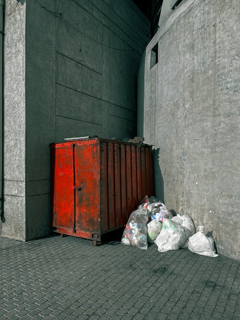 A weathered red metal waste container positioned against a dark, textured wall on the left and a lighter concrete wall on the right, situated on a paved urban sidewalk. Several white plastic rubbish bags, some partially open revealing mixed waste such as plastic bottles and packaging, are piled around the base of the container. The scene is outdoors, with natural ambient light illuminating the area, and the environment suggests an alleyway or backstreet setting typical for private rubbish collection or on-site clearance services. The overall appearance indicates a temporary waste storage area in need of removal, with a focus on independent rubbish disposal options that avoid local authority collection fees, aligning with the context of alternative waste handling available through professional rubbish clearance providers like Rubbish Clearance Bayswater.