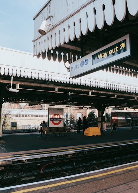 The image shows a train station platform beneath an overhanging structure with scalloped white trim and black support beams. A prominent directional sign hangs from the ceiling, indicating the lift and way out with arrows. In the background, there are a few people standing near the edge of the platform, with some sitting on benches. Behind them, the railway tracks are visible, with a partially visible station sign displaying the Golders Green station name. The environment appears to be outdoors, with natural daylight illuminating the scene. The platform surface is paved, and some yellow safety markings are visible along the edge. The scene conveys a typical urban train station setting with elements relevant to passenger movement and platform infrastructure, subtly reflecting services related to independent or private transportation and travel arrangements, in line with the context of rubbish removal and waste management services.