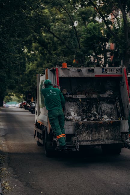 A worker in green uniform and high-visibility orange-striped trousers is loading or inspecting rubbish inside a large, white and red commercial waste collection vehicle parked on a quiet urban street. The vehicle's rear compactor area shows signs of previous use, with darkened, textured surfaces and residual debris. The scene is set amid clusters of trees with lush green foliage overhead, creating a shaded environment, while a few parked cars and the edge of a building are visible in the background. The worker appears to be engaged with the loading process, suggesting an active rubbish removal operation, which highlights the service offerings of independent waste collection providers such as Rubbish Clearance Bayswater. The overall atmosphere is calm, with natural lighting filtering through the trees, emphasizing the practicality of onsite or private waste disposal methods as an alternative to council bin collection, fitting within the context of efficient rubbish removal and clearance services.
