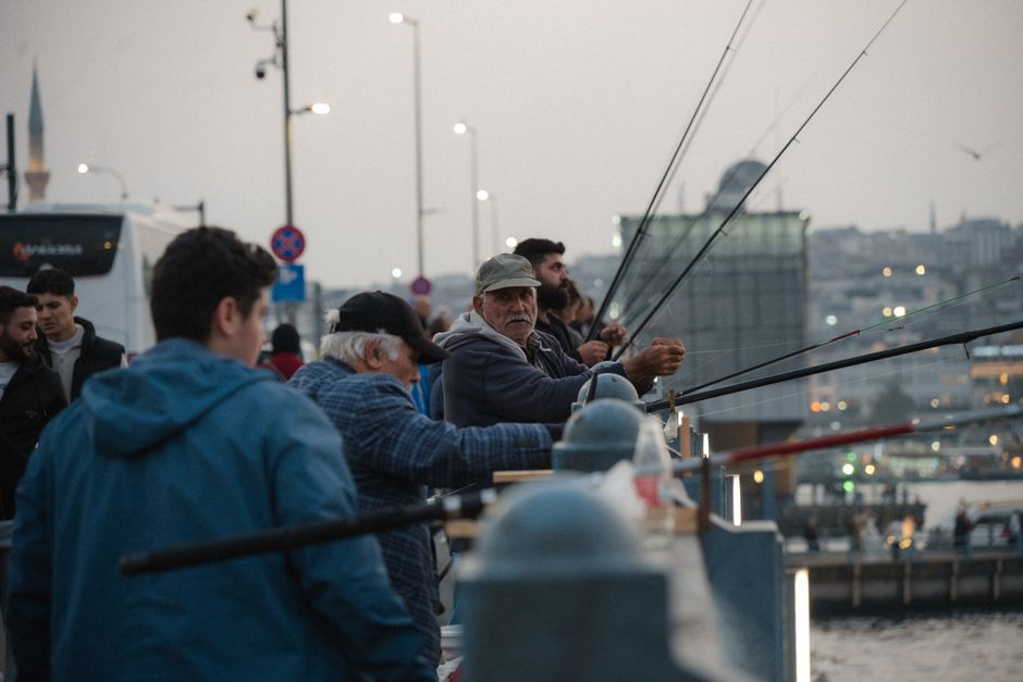 A group of men standing along a waterfront promenade during evening twilight, with some fishing rods in hand extended over the railing. The men are dressed in casual outerwear, such as jackets and caps, and appear to be engaging in fishing activity. The scene includes a background of cityscape with illuminated buildings, cranes, and boats moored in the water, suggesting an urban harbor environment. The sky is overcast with soft, diffused lighting, and streetlights along the promenade are lit. The overall atmosphere is calm and focused, reflecting a peaceful fishing activity possibly linked to private or independent waste handling if debris or waste associated with such activities is visible in the vicinity. Rubbish Clearance Bayswater offers solutions for waste removal from diverse settings, including outdoor recreational areas, which may be relevant here in an urban waterfront context.