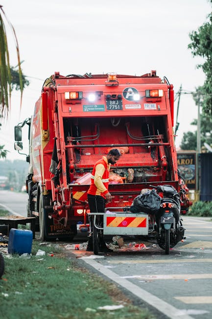 A worker in green uniform and high-visibility orange-striped trousers is loading or inspecting rubbish inside a large, white and red commercial waste collection vehicle parked on a quiet urban street. The vehicle's rear compactor area shows signs of previous use, with darkened, textured surfaces and residual debris. The scene is set amid clusters of trees with lush green foliage overhead, creating a shaded environment, while a few parked cars and the edge of a building are visible in the background. The worker appears to be engaged with the loading process, suggesting an active rubbish removal operation, which highlights the service offerings of independent waste collection providers such as Rubbish Clearance Bayswater. The overall atmosphere is calm, with natural lighting filtering through the trees, emphasizing the practicality of onsite or private waste disposal methods as an alternative to council bin collection, fitting within the context of efficient rubbish removal and clearance services.