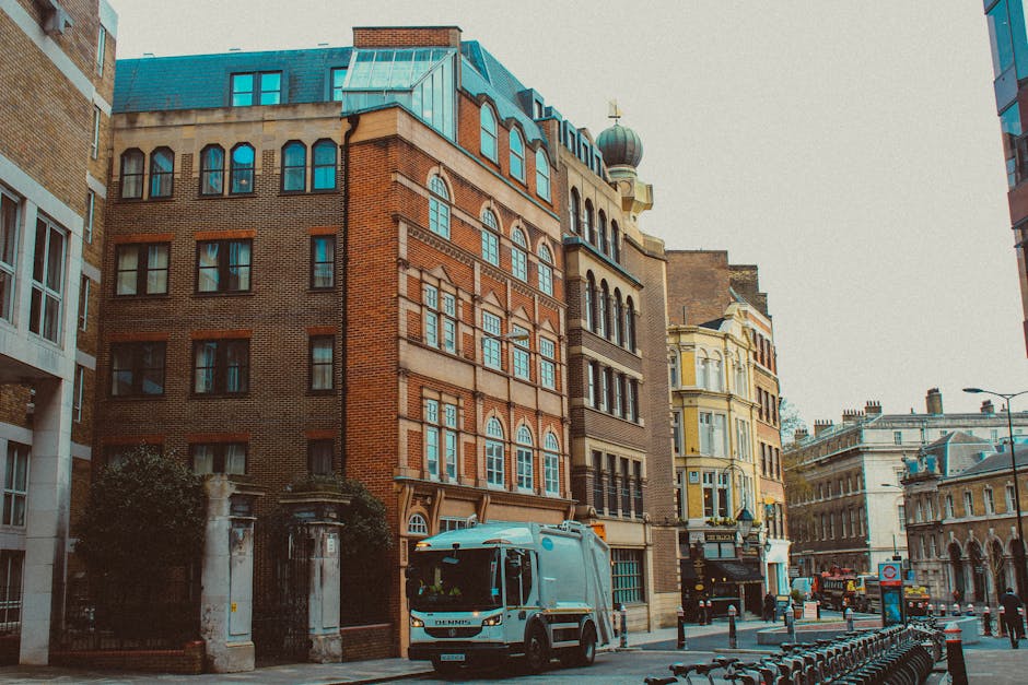 The image depicts a narrow urban street lined with multi-story buildings showcasing a variety of architectural styles, including brick facades and ornate window frames. A small refuse collection truck, painted light blue, is parked on the street close to the curb, suggesting active rubbish removal or collection service. The truck’s cabin features a modern design with clear windows and visible side mirrors, indicating it is used for commercial waste disposal. Surrounding the vehicle are metal bike racks positioned along the sidewalk, and in the background, other vehicles and street furniture are visible, including lampposts and a street sign. The lighting appears to be natural daylight with a slightly overcast sky, producing a neutral, even light across the scene. The street environment, with its tidy appearance and presence of waste collection equipment, subtly reflects the context of independent rubbish collection and waste management services, characteristic of private disposal sectors such as those provided by Rubbish Clearance Bayswater in the Bayswater area, supporting alternative rubbish removal options outside of local authority services.