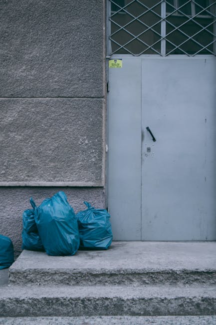 In front of a grey concrete building facade, four blue rubbish bags made of plastic are placed on a concrete pavement step, positioned to the left of a tall grey metal door with a black handle and a small windowed grid panel above it. The bags appear to be filled with waste and are loosely gathered with some wrinkles and creases visible on their surfaces. The building wall, with its textured, rough finish, extends vertically on the left, while the metal door to the right features a slightly weathered surface and is part of a larger metal frame grid above it. The scene is illuminated by natural daylight, casting soft shadows, and the overall setting suggests an urban area set for waste collection or private rubbish removal, with the presence of the waste bags indicating a small-scale rubbish clearance effort managed by a service such as Rubbish Clearance Bayswater. The environment exemplifies typical private waste handling outside a commercial or residential building, aligning with non-local authority rubbish disposal practices often seen in independent rubbish removal services.