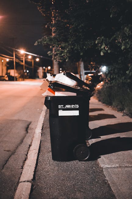 A black wheelie bin labeled 'St. John's' sits on a residential sidewalk at night, filled with various types of rubbish including cardboard boxes and plastic bags that are overflowing from the open lid. The bin appears to be made of durable plastic with a textured surface, and the white label and logo are clearly visible. Positioned on a paved driveway or pavement, the bin casts a shadow on the ground due to nearby street lighting. In the background, there are trees with dark foliage, and a quiet street illuminated by orange-hued streetlights. The surrounding environment suggests an urban or suburban area where residents manage their waste collection privately, perhaps reflecting on alternative rubbish disposal methods in keeping with local regulations or community standards. This scene emphasizes the importance of appropriate rubbish storage and disposal practices for effective waste management, consistent with services provided by companies like Rubbish Clearance Bayswater for rubbish removal and on-site clearance needs.
